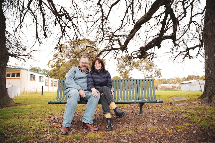 A man in a knit sweater and jeans sits on a park bench with his arm around a woman wearing jeans and a puffer jacket, smiling.
