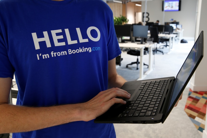 A male employee wearing a Booking.com-branded T-shirt stands and uses a laptop with one hand inside an office.
