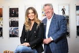 Delta Goodrem and Denis Handlin smile standing in a room with pictures of musicians on the wall. Delta is leaning on a table.