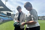 Two women stand on a cricket ground looking at a green cap.