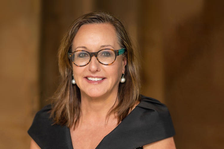 A woman with glasses, pearl earrings and long fair hair smiles.