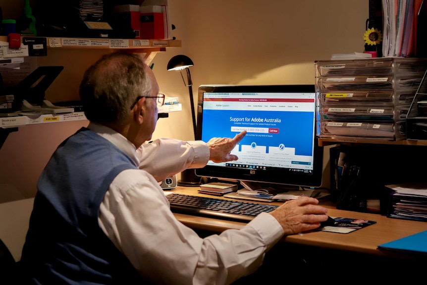 Geoff Sussman, wearing a white shirt and blue vest, points at his computer in his study. The Adobe support page is on screen