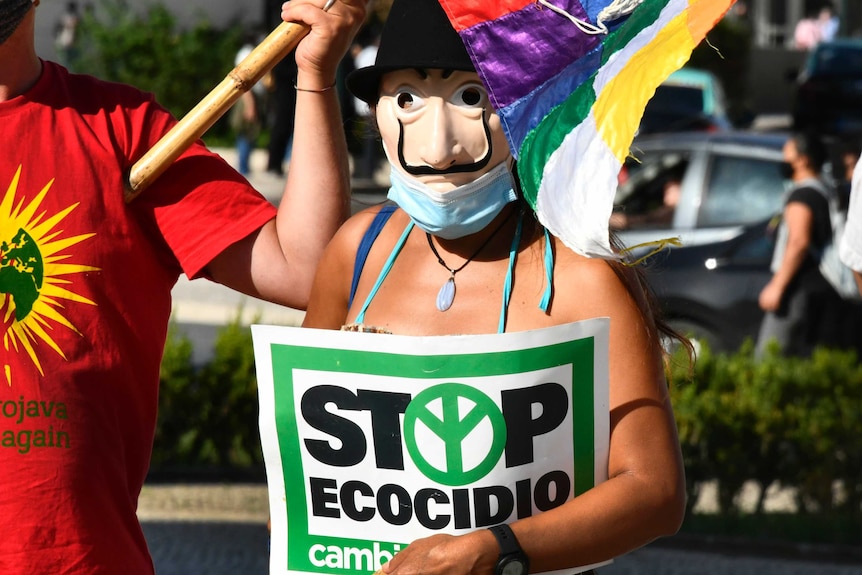 A climate change activist wearing a mask holds a sign which reads "Stop Ecocide".