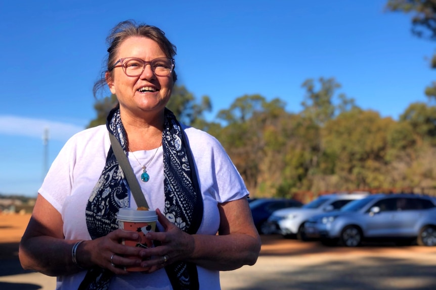 A woman wearing glasses and a white t-shirt stands holding a takeaway coffee cup in a carpark.