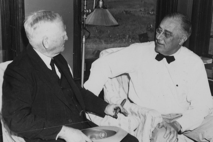 A black and white photo of FDR in a white tuxedo talking to a man in a black suit 