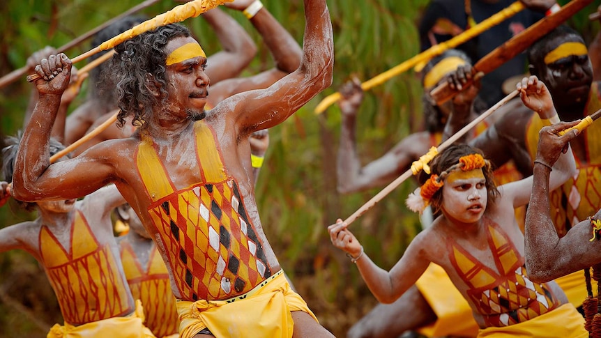 Dancers in traditional body paint holding sticks wrapped in material above their heads.