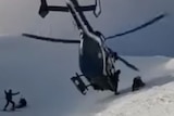 a man holds up his hands as a helicopter lands on a mountain next to a man lying down