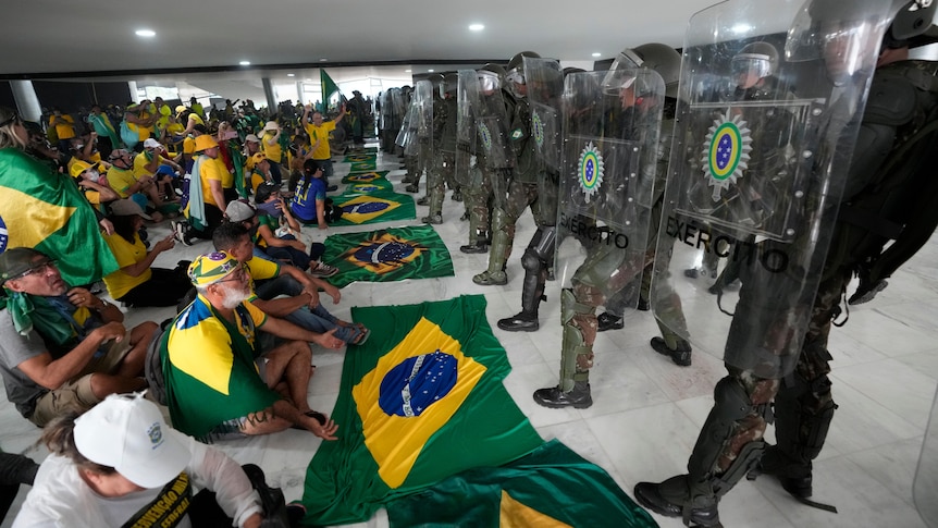 Protesters sit on the ground with a row of Brazilian flags in front of riot police.