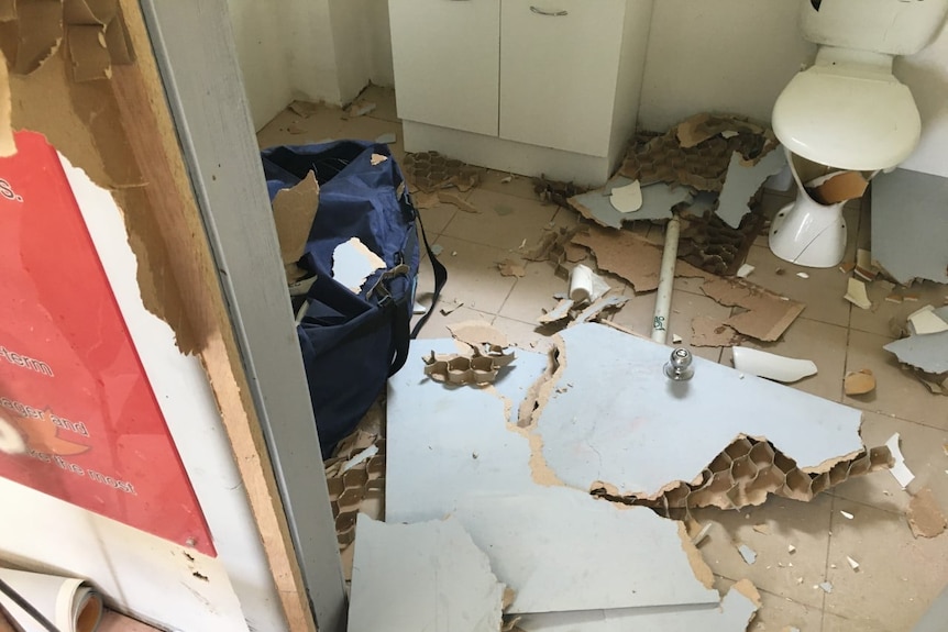 A bathroom littered with debris. The toilet is smashed and pipes have been torn out of their fixtures.