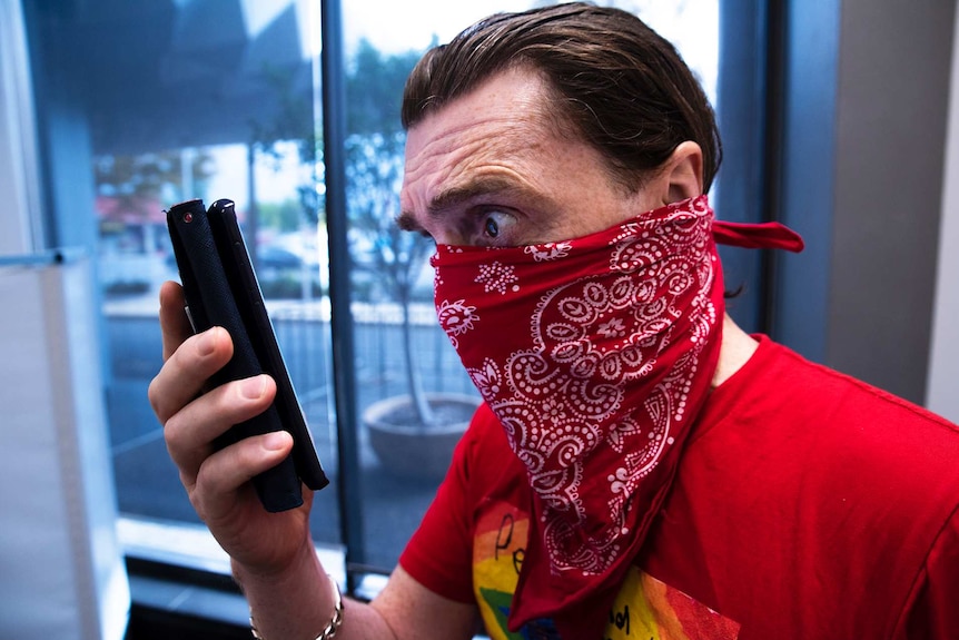 A man with a handkerchief over his mouth and nose looks fearfully at a smart phone.