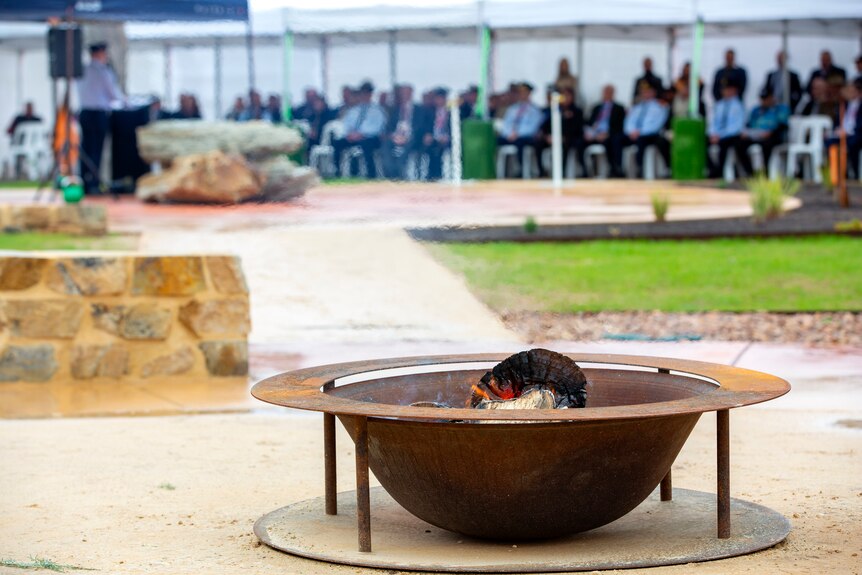 A metal fire pit with people seated for an address in the background