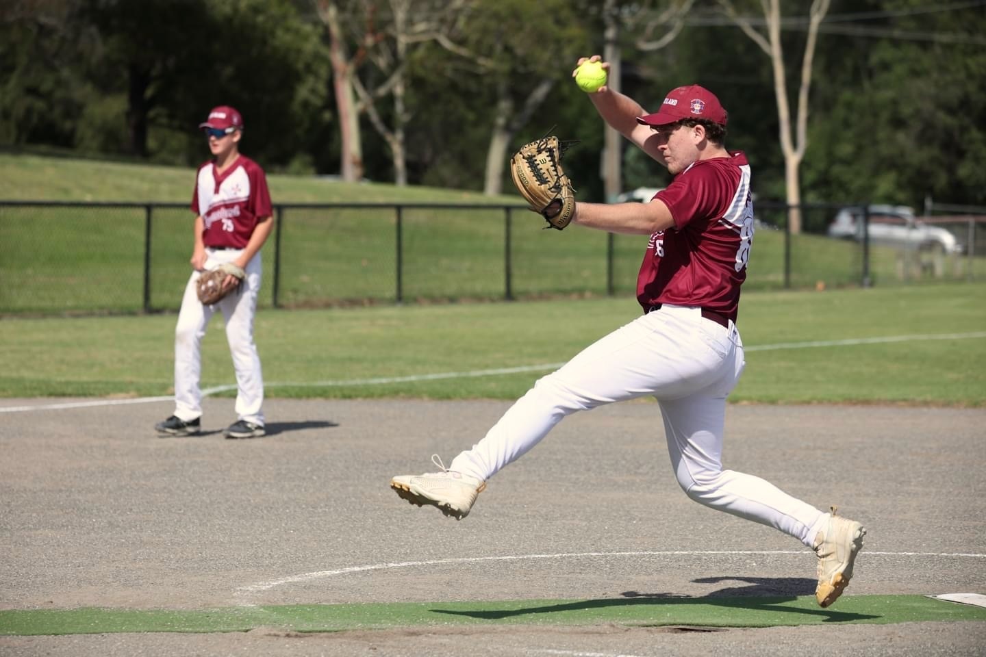 A teenage boy wearing a maroon and white softball uniform and brown softball glove strides out to pitch a yellow softball.