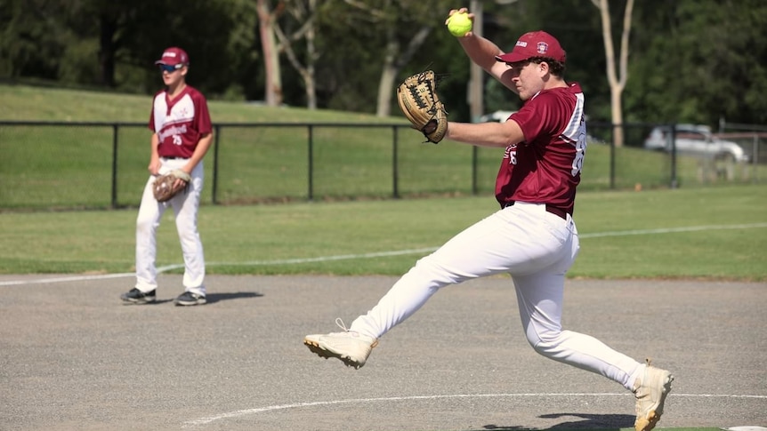A teenage boy wearing a maroon and white softball uniform and brown softball glove strides out to pitch a yellow softball.