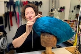 A woman makes adjustments to on a blue feathered hat on a wooden dummy.