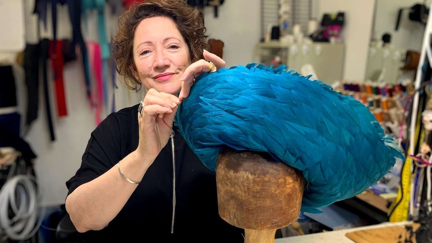 A woman makes adjustments to on a blue feathered hat on a wooden dummy.