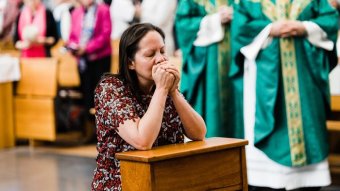 A woman kneels with her eyes closed and hands cupped to her mouth in prayer with blurred priest robes in background.