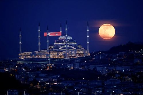 Bright yellow full moon rising to the right of a large mosque with red flag f lying  with white crescent moon and star on it