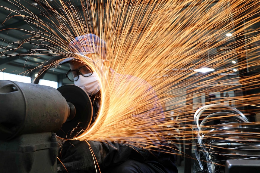 Sparks coming off a manufacturing machine while a factory worker in a mask looks on