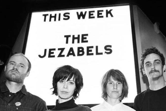 Black and white photograph of four people with a neon sign behind them that says This Week The Jezabels