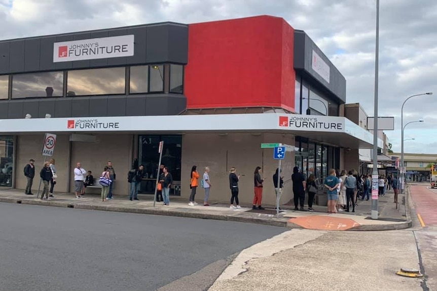 People stand in a line outside a building, with the queue extending around a block of shops