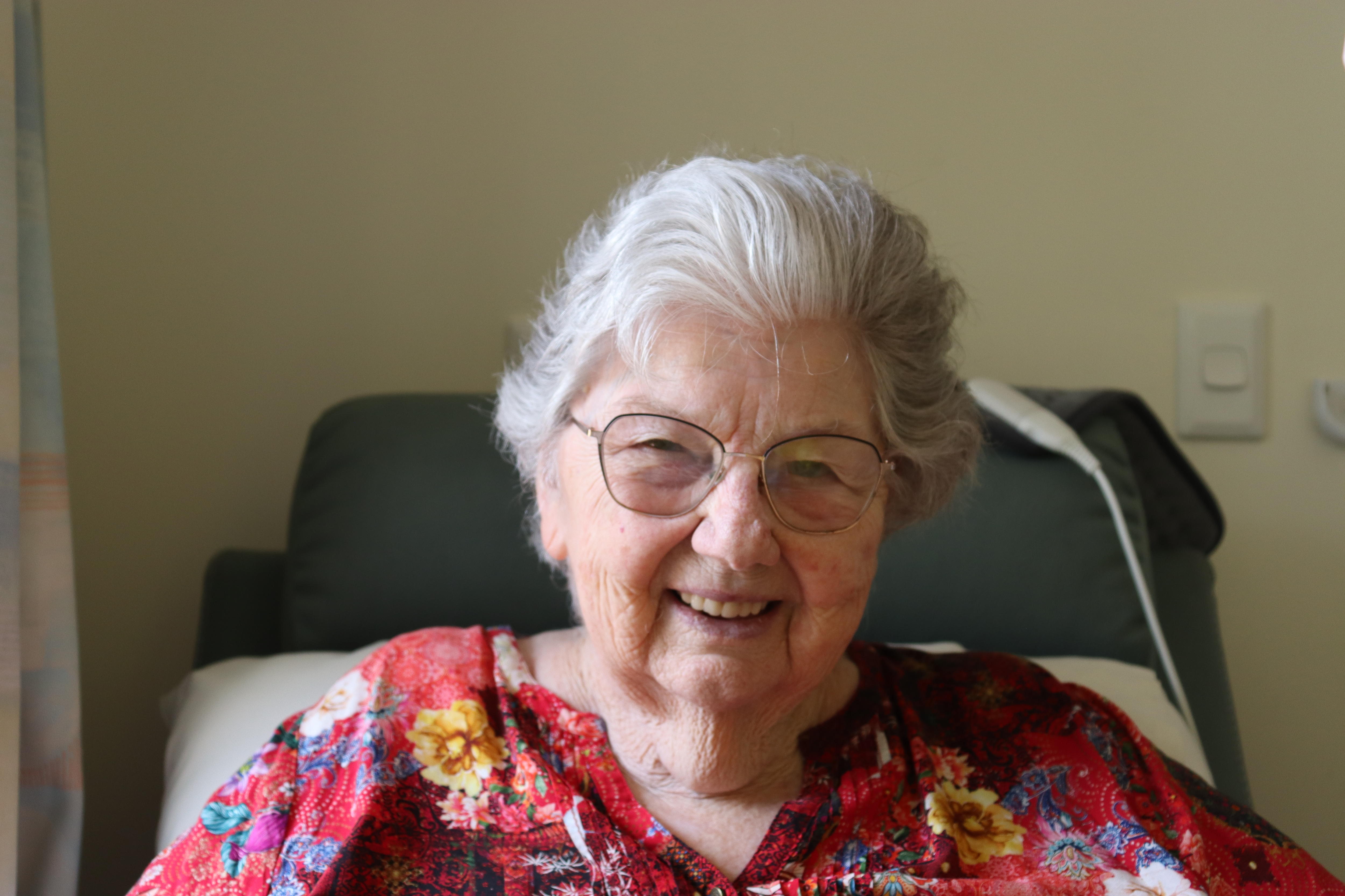 An elderly woman wearing glasses and a bright red floral shirt smiles, she is sitting up in an aged care bed