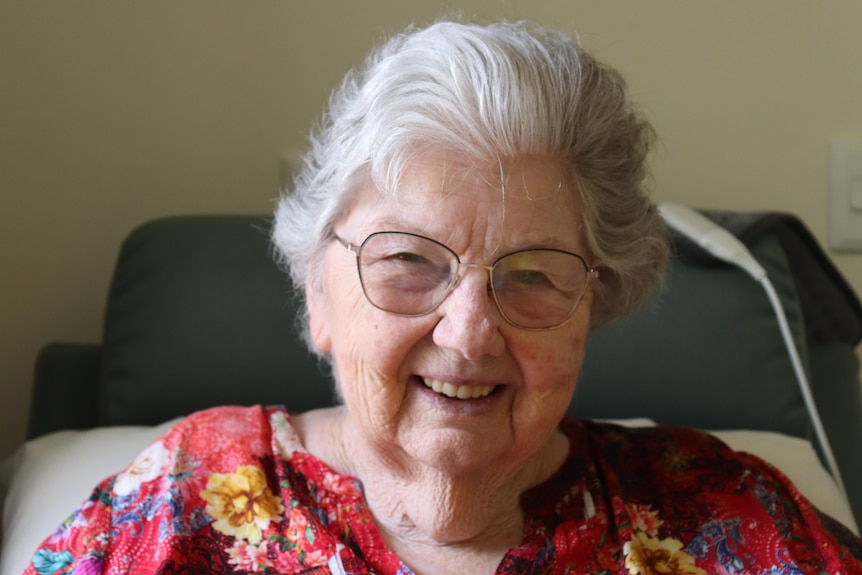 An elderly woman wearing glasses and a bright red floral shirt smiles, she is sitting up in an aged care bed
