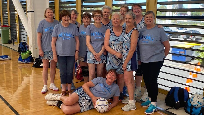 12 women some in blue netball dresses and others in blue t-shirts stand on the side of a netball court