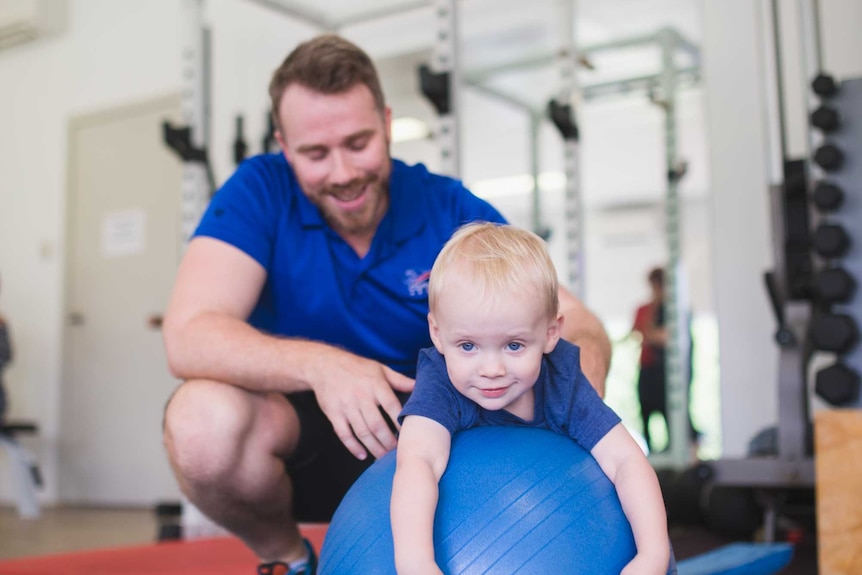A physiotherapist squats behind an exercise ball, where a toddler lays laughing