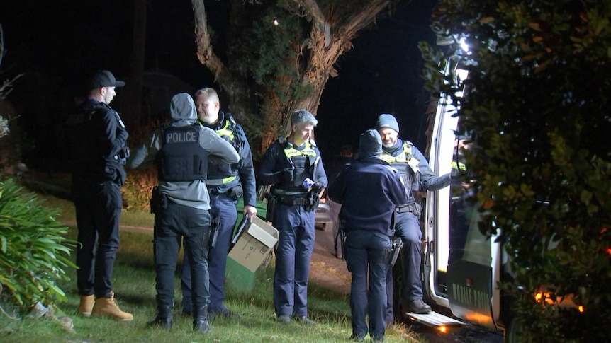 Police officers stand about in the darkness outside a police van.