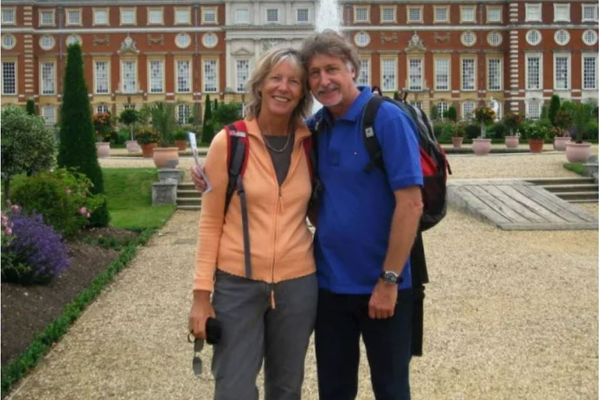 A man wearing a blue shirt and backpack stands with a woman wearing a backpack in front of a fountain and building. 