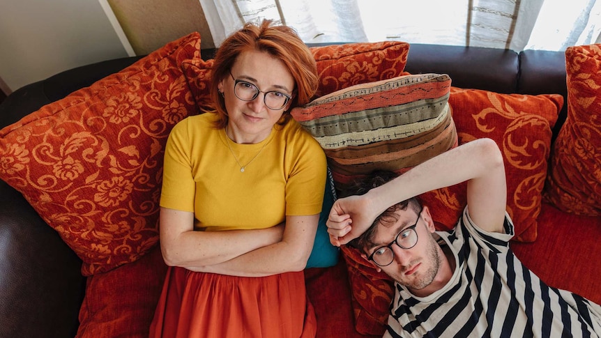 A mother and her grown son sitting on a couch with lots of rusty coloured cushions