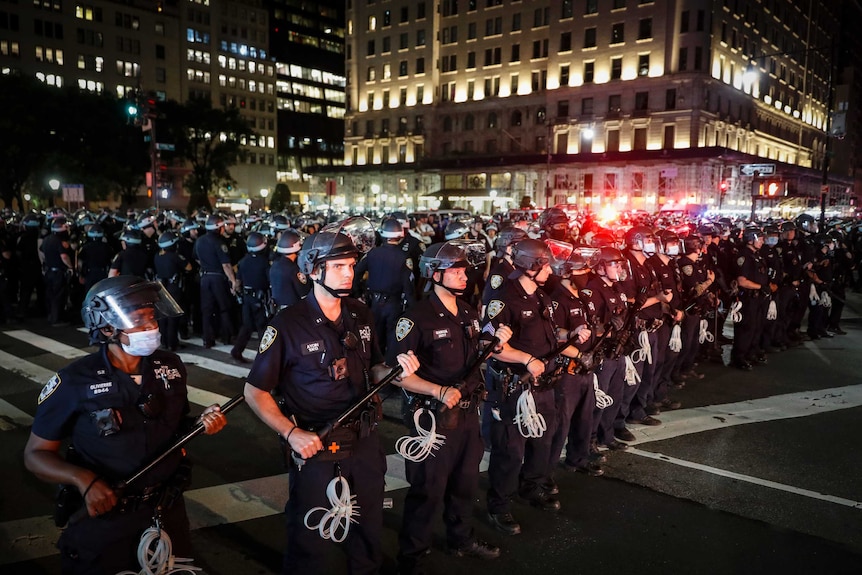 Hundreds of uniformed police wearing riot helmets and carrying truncheons form up in a line facing protesters.