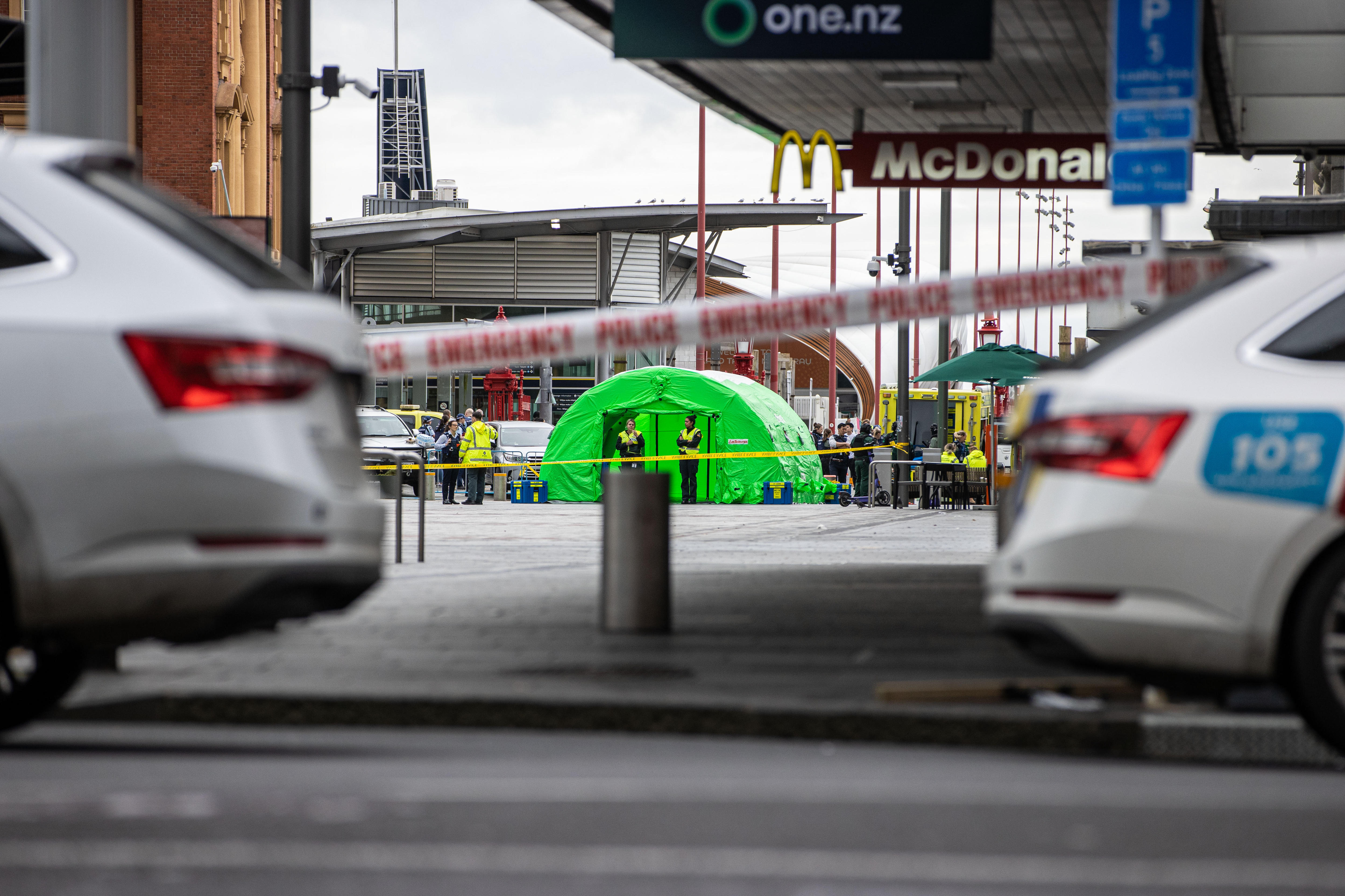 A green tent among a crowd of paramedics wearing high vis vests near a constrction site. police cars and tape in the foreground