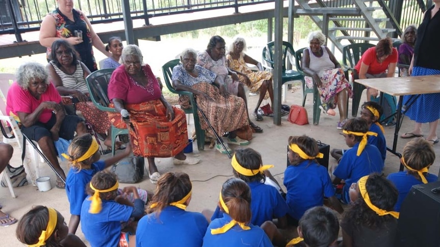 A group of Aboriginal elder women sitting on chairs, speaking to students who sit on the floor in front of them 