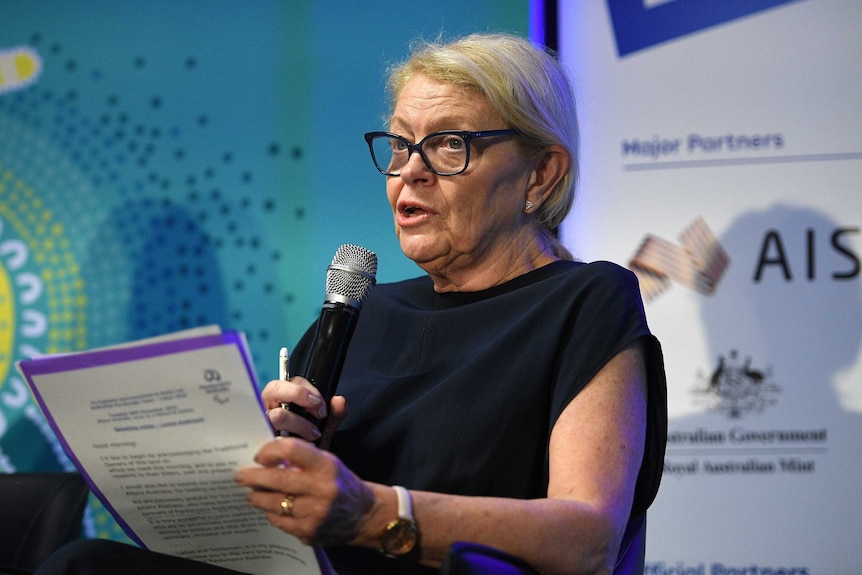 Lynne Anderson, wearing black-rimmed glasses and a black dress, holds a microphone in one hand and a piece of blue-trimmed paper