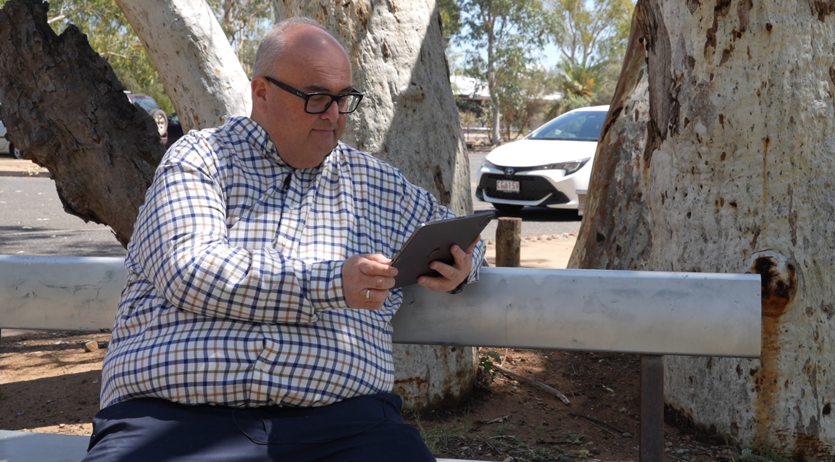 Danial Rochford sits on a park bench in front of a native Australian tree looking at an electronic tablet.