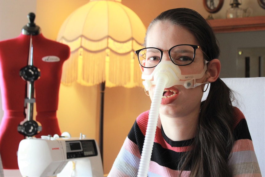 A profile shot of Amy Evans on a ventilator and wearing spectacles posing for a photo, in front of a lamp and sewing machine.