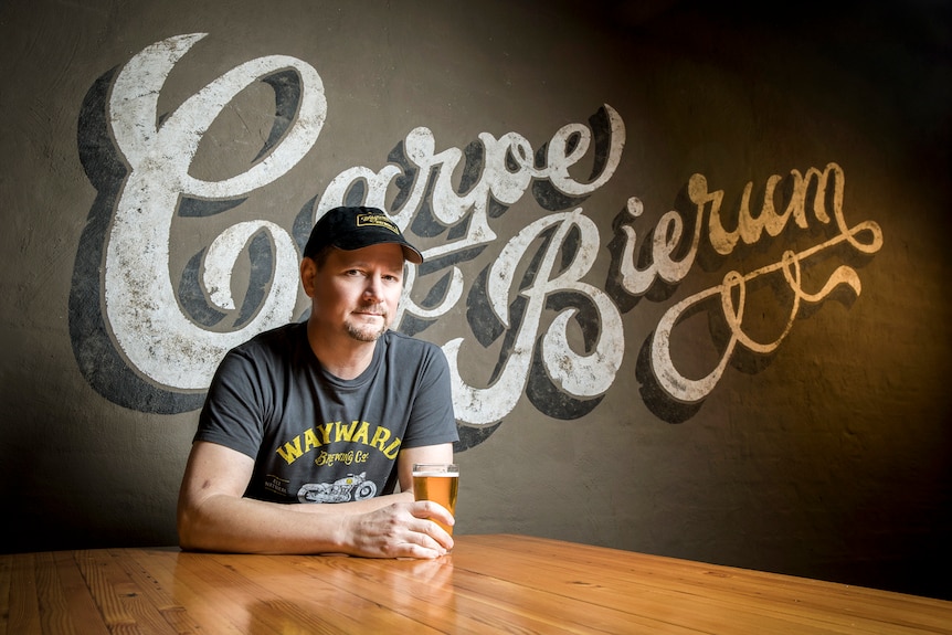 A man wearing a hat sits at a wooden table holding a glass of beer