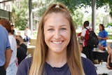 A young woman with long blonde hair smiles at camera in front of group of students in background.