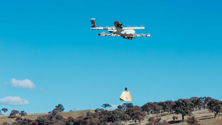 A a cardboard box hangs from a multi-winged drone that is flying through the sky.