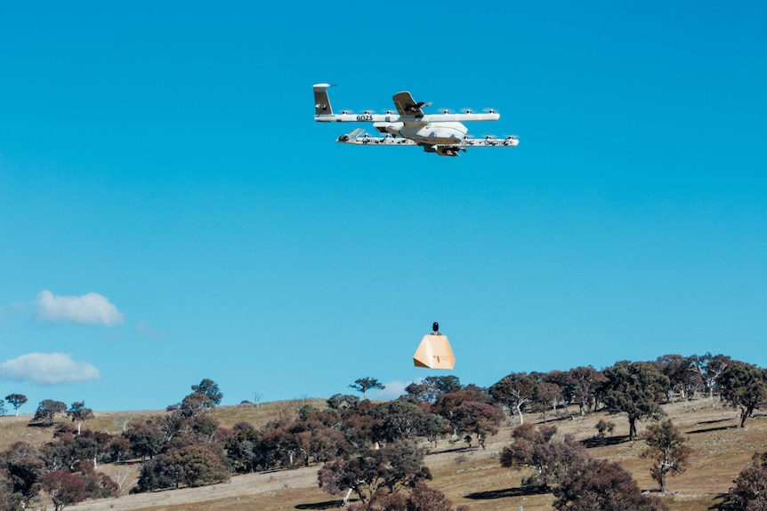 A a cardboard box hangs from a multi-winged drone that is flying through the sky.