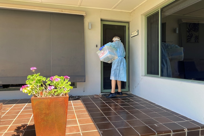 Photo shows back of nurse in full PPE standing at a front door with a plastic tub under one arm.