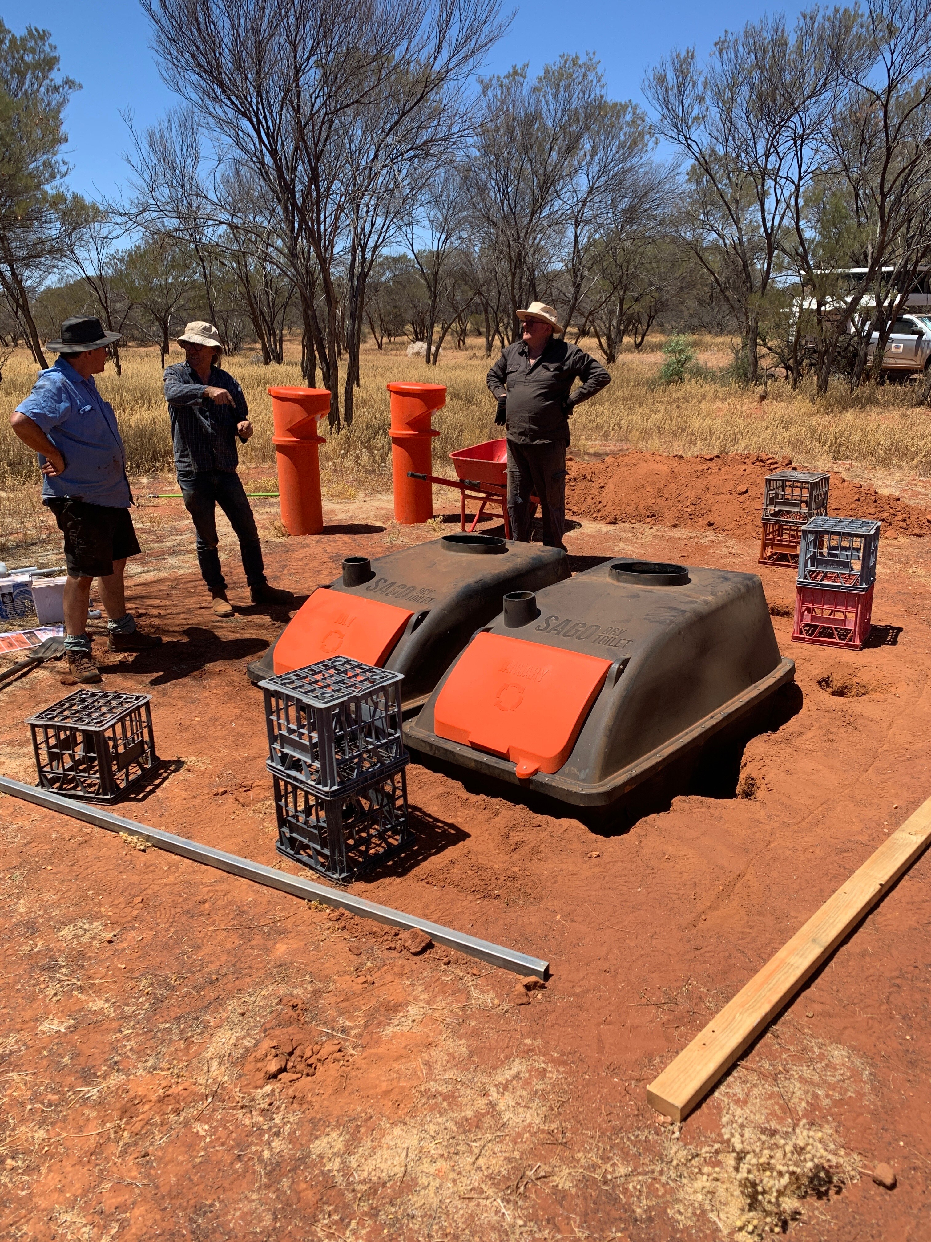 Three men wearing hats have a conversation while standing on red dirt