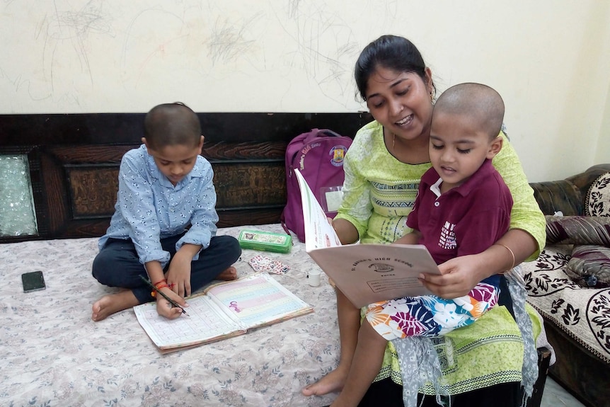 A woman wearing a yellow shirt holds a boy on her lap as she reads a book with another boy sitting beside her.