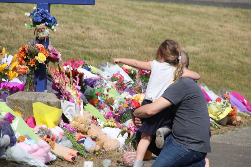 A small girl wraps her arm around the shoulders of her father in front of flower tributes.