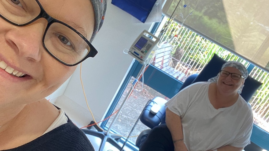 Two women in a hospital ward wearing bandanas smile for a selfie.