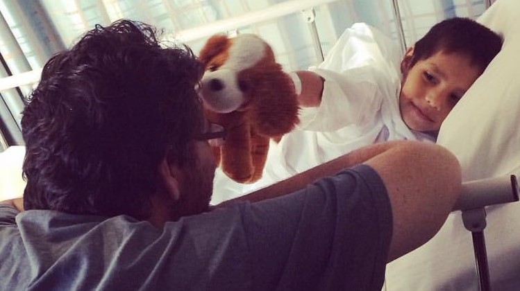 A boy in a hospital bed with smiles and plays with stuffed animal with his dad.