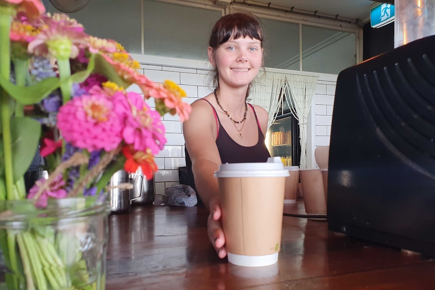 A smiling young woman serves a cup of coffee in a cardboard cup with flowers in the foreground and a brick wall behind