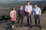 Four young medical students in professional clothing stand on a high bushland ridge with town and trees receding behind them