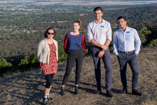 Four young medical students in professional clothing stand on a high bushland ridge with town and trees receding behind them